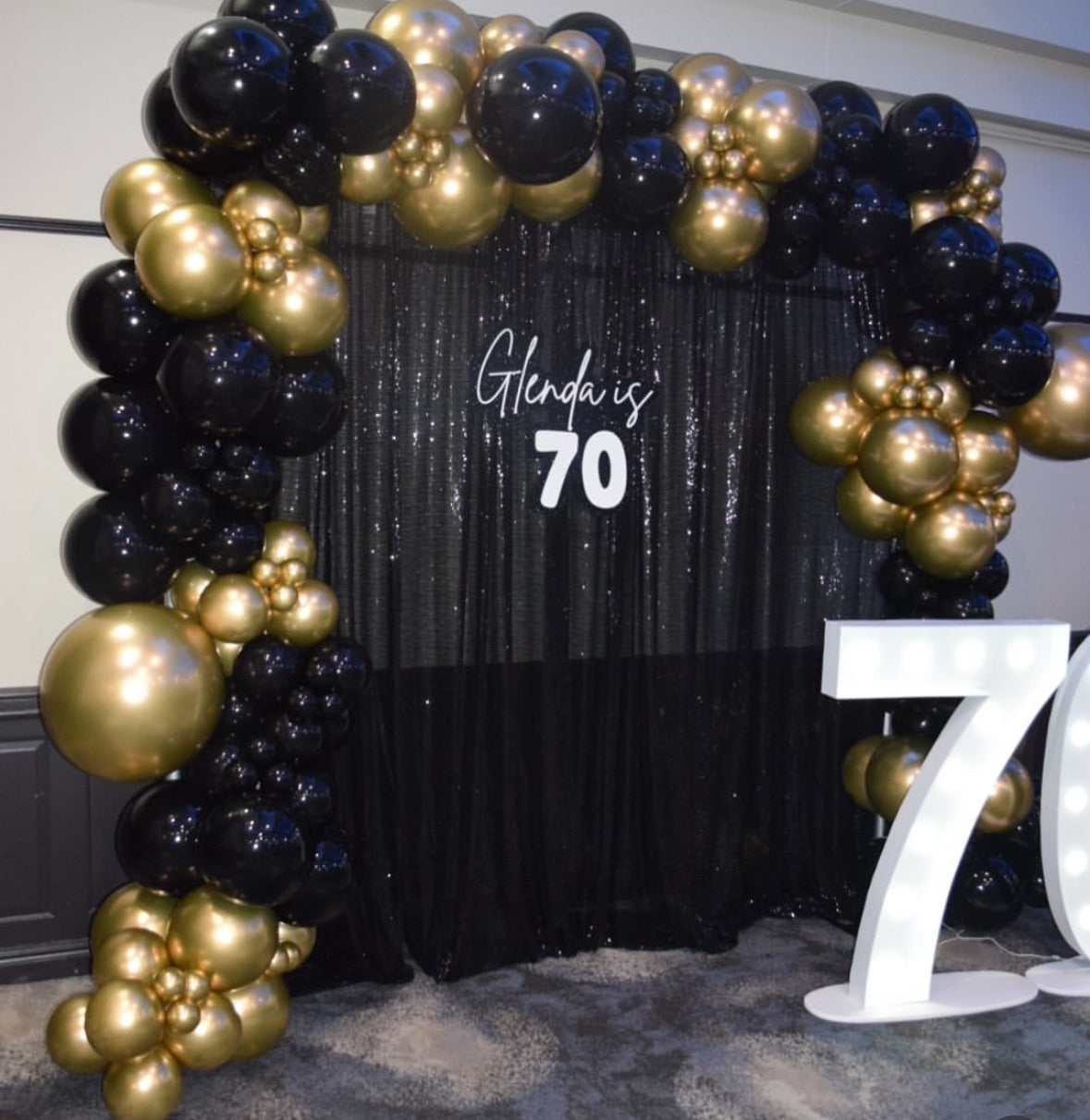 Black shimmer curtain with 'Glenda is 70' sign and balloon arch around it